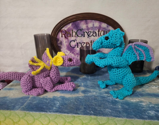 Two crocheted dragons, one purple and one blue, on a textured surface with a 'RahCreatives' sign in the background.