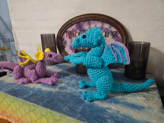 Two crocheted dragons, one blue and one purple, on a table with a framed picture in the background.