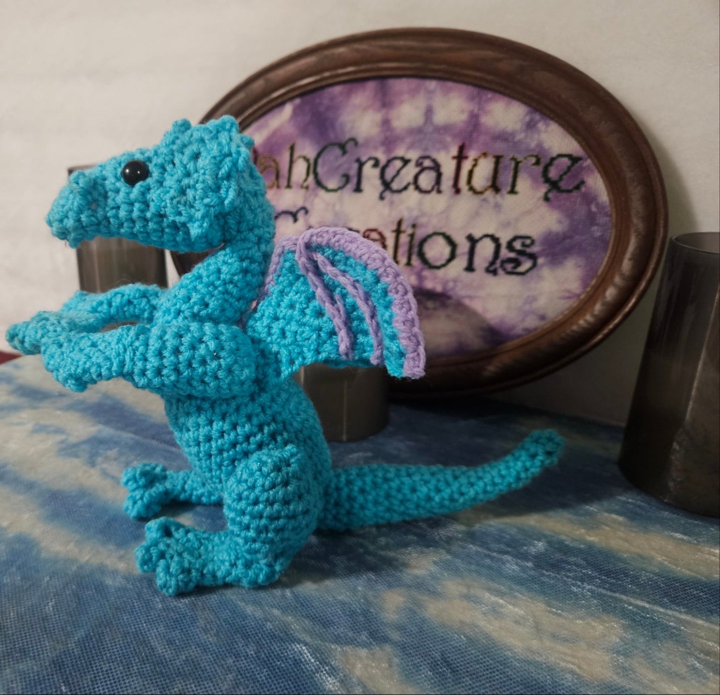 Crocheted blue dragon toy on a textured surface with 'Fable Creature Creations' sign in the background.