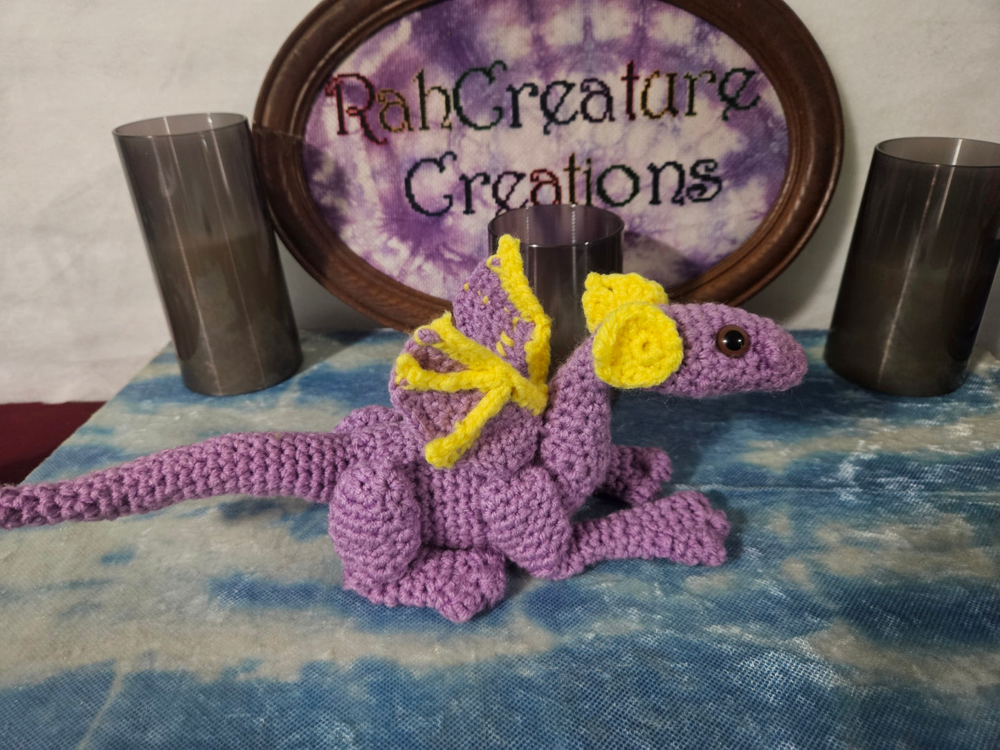 Purple crocheted creature with yellow accents on a textured surface with 'RobCreature Creations' sign in the background.