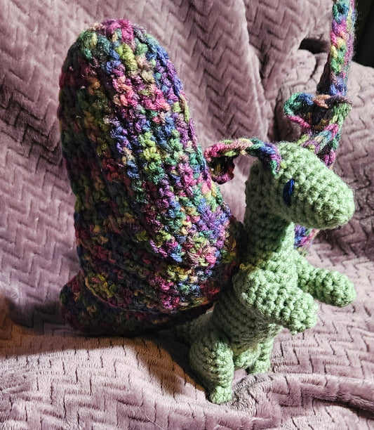 A handmade crocheted snail toy with a green body and a multicolor shell, resting on a textured pink blanket.
