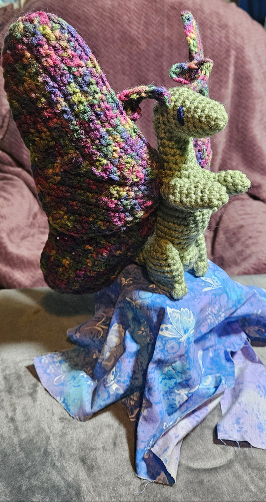 A handmade crocheted dragon figurine with multicolored wings and a green body, posed on a blue patterned fabric base in a cluttered craft room setting.