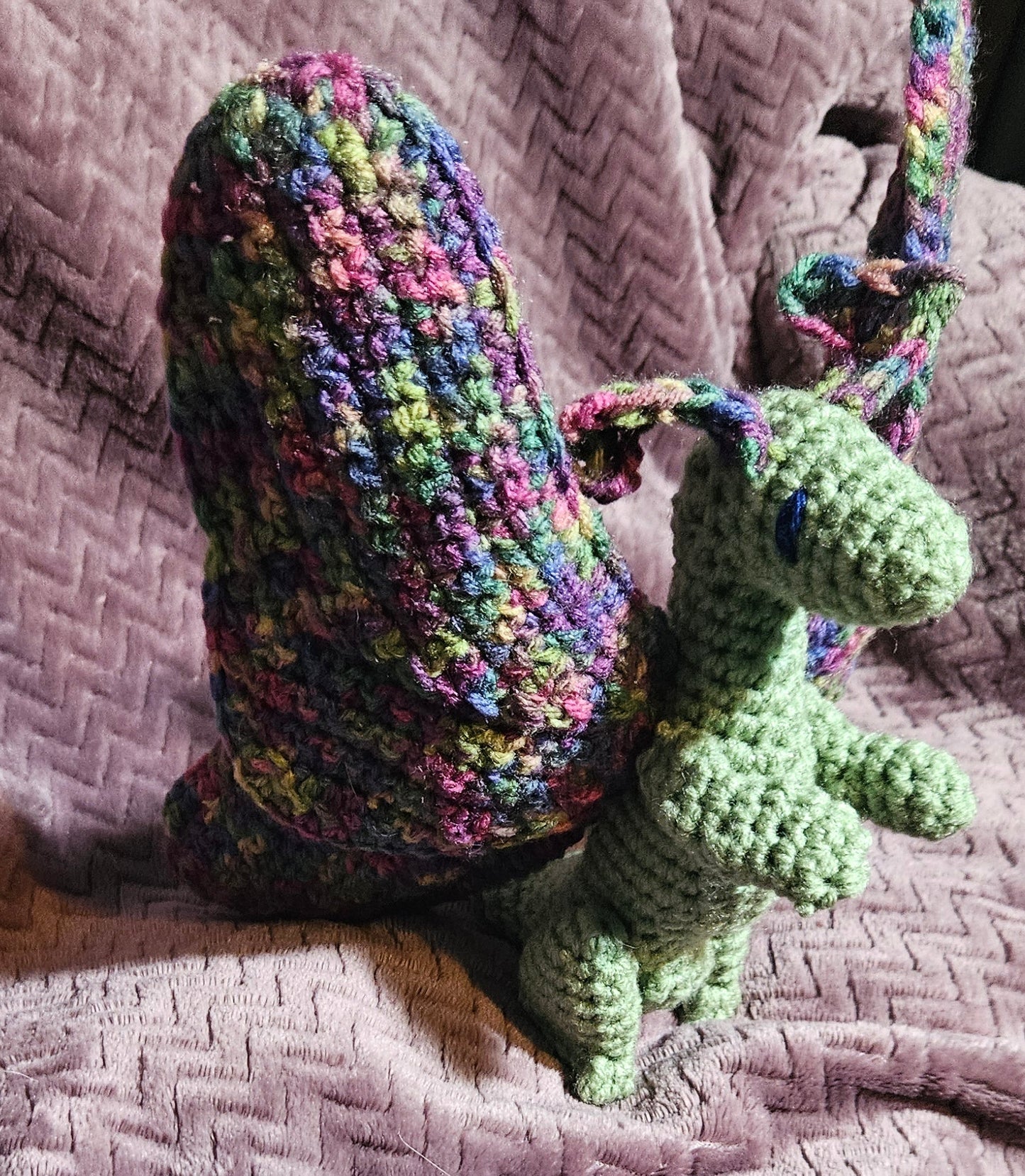 A handmade crocheted snail toy with a green body and a multicolor shell, resting on a textured pink blanket.