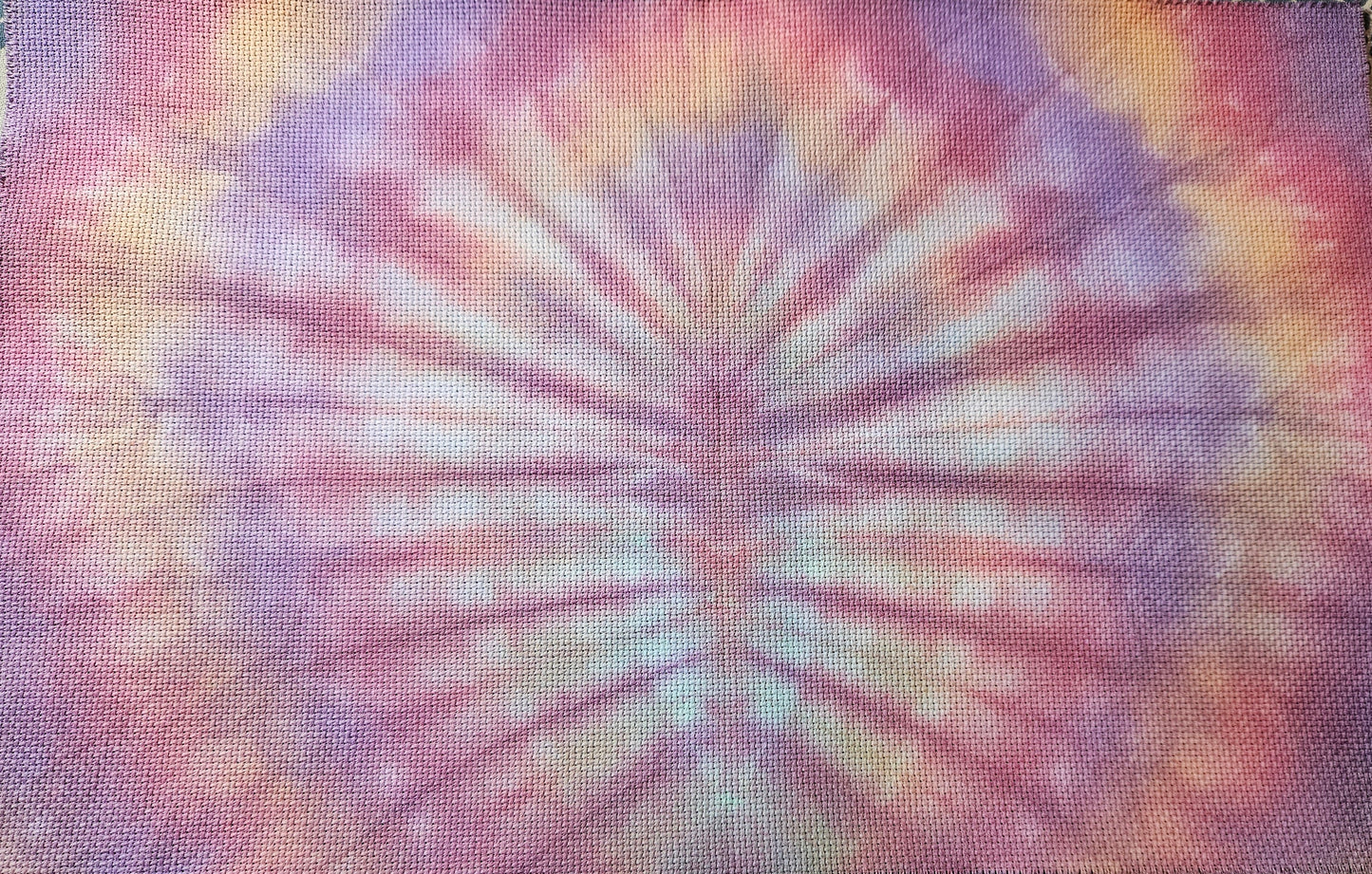 A square piece of textured fabric with a vibrant tie-dye pattern featuring purple, pink, orange, and yellow rays radiating from the center, laid flat on a patterned surface.