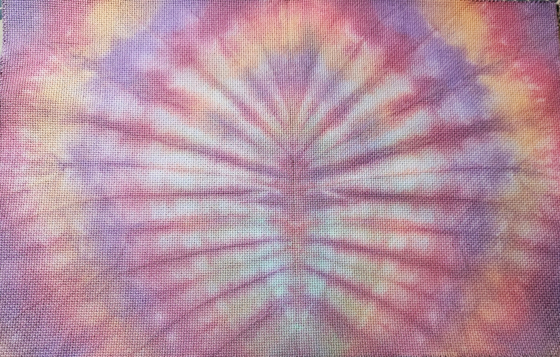 A square piece of textured fabric with a vibrant tie-dye pattern featuring purple, pink, orange, and yellow rays radiating from the center, laid flat on a patterned surface.