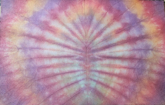 A square piece of textured fabric with a vibrant tie-dye pattern featuring purple, pink, orange, and yellow rays radiating from the center, laid flat on a patterned surface.
