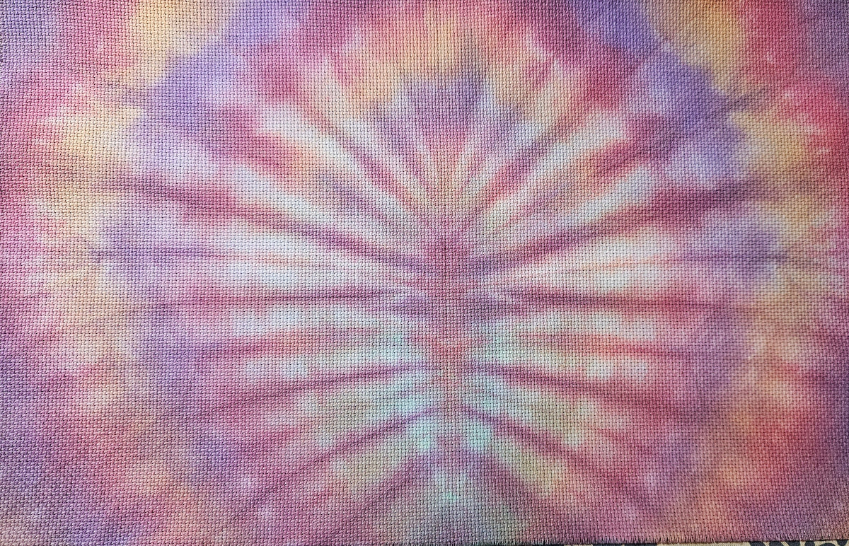 A square fabric textile with a vibrant tie-dye spiral pattern in pink, purple, orange, and white, laid flat on a patterned fabric background.