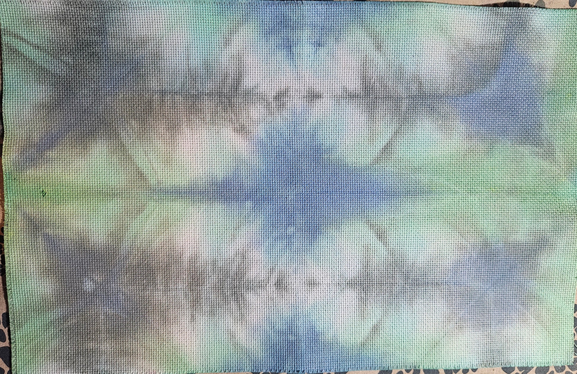 A rectangular piece of fabric with a blue, green, and white tie-dye pattern, laid flat on a patterned surface.